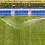 Luftbild eines Fußball-Vereinsplatzes: Großregner beregnen den Rasen vor der Zuschauertribüne